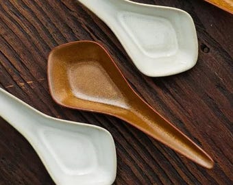 May include: Four ceramic spoons, two brown and two white, are arranged on a wooden surface. The spoons are all slightly curved and have a rounded bowl.