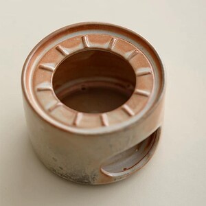 May include: A cylindrical ceramic incense burner with a circular opening at the top and a smaller opening on the side. The burner is primarily beige with reddish-brown accents around the top rim and interior.