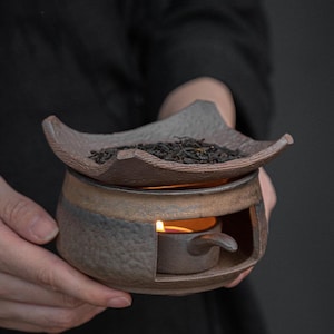 May include: A ceramic tea warmer with a candle inside, topped with a tray of loose tea leaves. The warmer has a brown and gray color scheme, with a small handle on the side. The tea warmer is designed to keep tea warm.