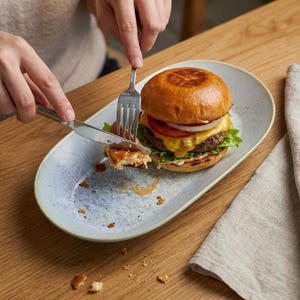 May include: A person using a fork and knife to cut a burger on a light blue oval plate. The burger has a sesame seed bun, a beef patty, cheese, tomato, lettuce, and onion. A beige linen napkin is on the right.