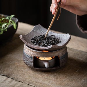 May include: A ceramic tea warmer with a square tray holding loose tea leaves. A small candle burns inside the warmer, and a person is using wooden tongs to handle the tea. The warmer has a rustic, textured finish.