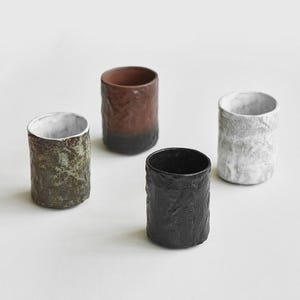 Puede incluir: Cuatro vasos de cerámica hechos a mano con superficies texturizadas. Los colores incluyen un marrón oscuro con base negra, un vaso negro, un vaso con estampado verde y marrón y un vaso blanco y gris. Los vasos tienen una estética rústica y orgánica.