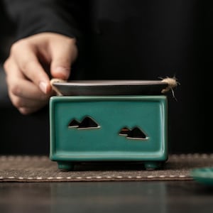 Op de afbeelding: Een turquoise keramische wierookbrander met een rechthoekige basis en bergvormige uitsparingen. Bovenop ligt een kleine, donkere lade, verbonden met een natuurlijke vezelkoord. De brander is ontworpen voor wierookstokjes of -kegels.