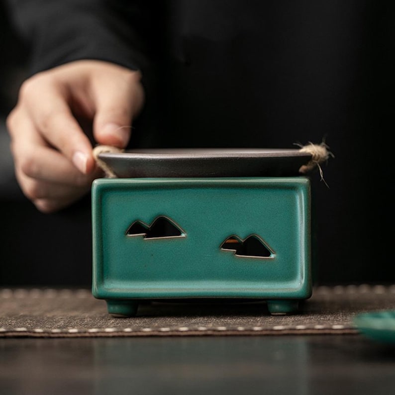 Op de afbeelding: Een turquoise keramische wierookbrander met een rechthoekige basis en bergvormige uitsparingen. Bovenop ligt een kleine, donkere lade, verbonden met een natuurlijke vezelkoord. De brander is ontworpen voor wierookstokjes of -kegels.