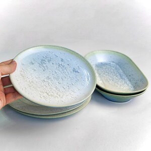 May include: A set of ceramic plates and bowls with a speckled, gradient design. The plates are round, and the bowls are oval-shaped. The color palette includes shades of blue, white, and green. The plates are stacked, and one is being held.