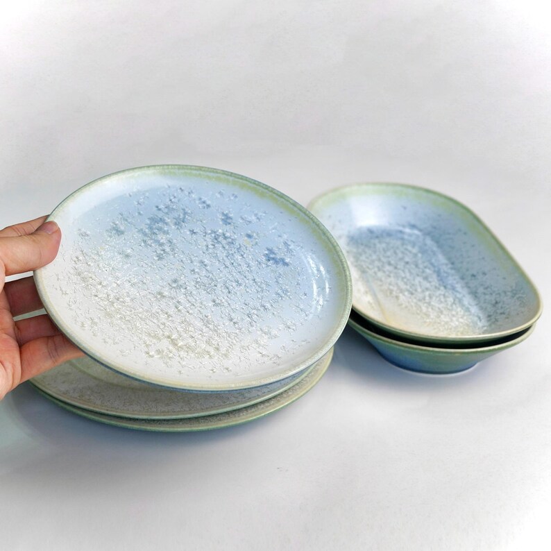May include: A set of ceramic plates and bowls with a speckled, gradient design. The plates are round, and the bowls are oval-shaped. The color palette includes shades of blue, white, and green. The plates are stacked, and one is being held.