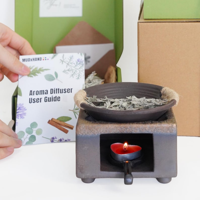 Op de afbeelding: Aromaverspreider met een donkerbruine keramische kom en een vierkante basis. De kom is gevuld met gedroogde kruiden en heeft een touwrand. Een brandende rode kaars staat eronder. De gebruikershandleiding luidt "Aroma Diffuser User Guide".