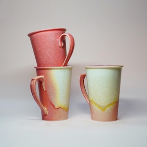 Puede incluir: Tres tazas de cerámica con un esmalte rosa y amarillo. Las tazas tienen una forma alta y cilíndrica y un asa en el lateral.