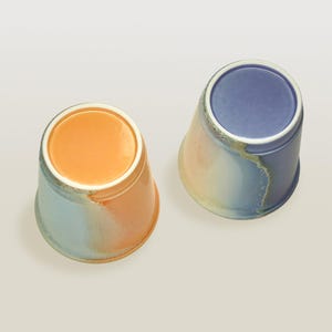 May include: Two ceramic cups with a gradient glaze. One cup is blue, white, and orange. The other cup is orange, white, and blue.