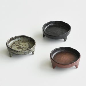 Puede incluir: Tres cuencos pequeños de cerámica hechos a mano con una estética rústica. Cada cuenco es redondo con un borde ligeramente irregular y se apoya en tres patas cortas. Los cuencos están esmaltados en tonos negro, marrón y verde, mostrando una superficie texturizada.