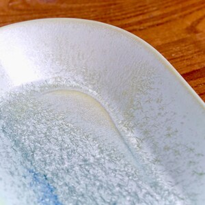 May include: Close-up of a white ceramic dish with a textured, crystalline pattern. The dish has a curved edge and a shallow, rounded interior. The surface has a subtle, iridescent sheen with hints of blue and yellow.