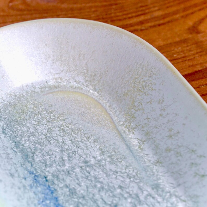 May include: Close-up of a white ceramic dish with a textured, crystalline pattern. The dish has a curved edge and a shallow, rounded interior. The surface has a subtle, iridescent sheen with hints of blue and yellow.