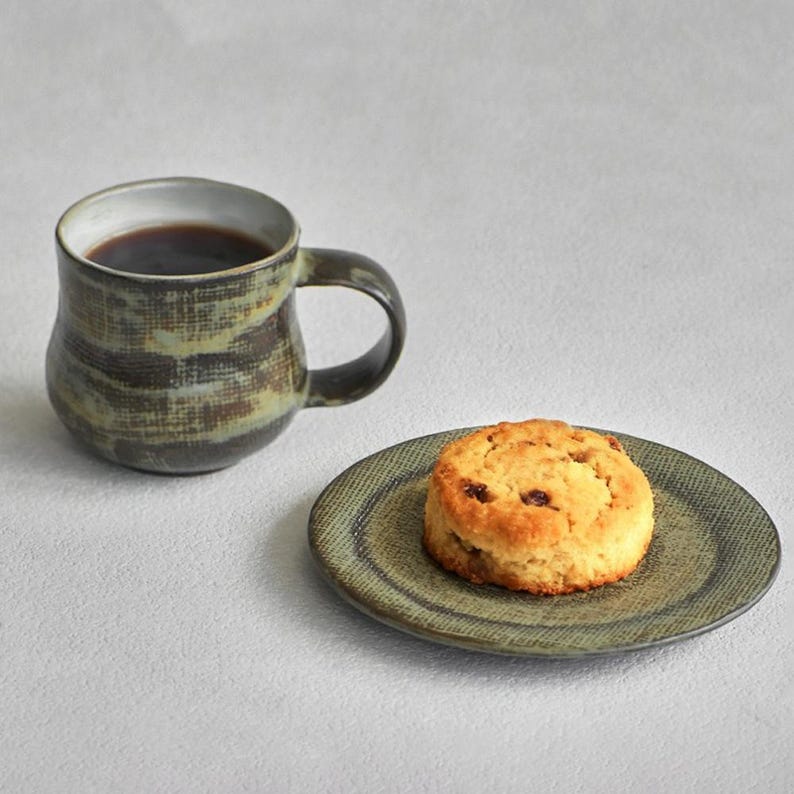 K&ouml;nnte beinhalten: Eine Keramiktasse gef&uuml;llt mit dunkler Fl&uuml;ssigkeit, neben einem kleinen Teller mit einem Scone. Die Tasse hat ein strukturiertes, erdiges Design in Braun- und Gr&uuml;nt&ouml;nen. Teller und Scone haben &auml;hnliche Erdt&ouml;ne.