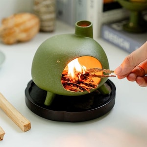 May include: A green ceramic incense burner with a spherical body, three small legs, and a chimney. The burner has a circular opening with burning incense inside. It sits on a dark, round tray.