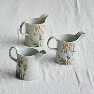 Op de afbeelding: Drie keramische kannen met bloemmotieven. De kannen zijn gebroken wit met handgeschilderde bloemen in geel, oranje en blauw. Elke kan heeft een handvat en een tuit. De kannen staan op een witte ondergrond.