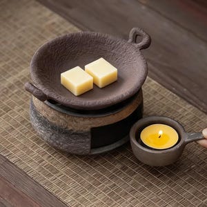 May include: A brown ceramic wax warmer with a small handle and a dish holding two yellow wax cubes. A lit tea light candle sits in a matching small cup with a handle. The warmer sits on a woven mat.