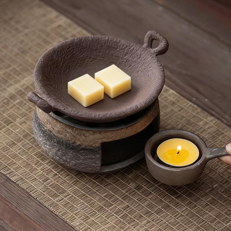 May include: A brown ceramic wax warmer with a small handle and a dish holding two yellow wax cubes. A lit tea light candle sits in a matching small cup with a handle. The warmer sits on a woven mat.