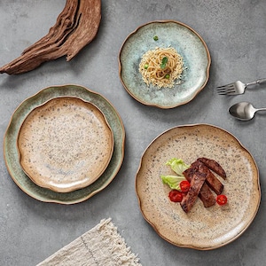 Puede incluir: Conjunto de platos de cerámica rústica con bordes irregulares. Los platos presentan una superficie moteada y texturizada en tonos marrones, verdes y azules. Algunos platos están llenos de comida, incluyendo pasta y carne.