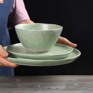 Op de afbeelding: Een driedelige keramische serviesset in een gespikkelde mintgroene glazuur. De set bevat een kom en twee borden, allemaal met een witte basis. De borden hebben verschillende maten. Het servies wordt door een persoon vastgehouden.
