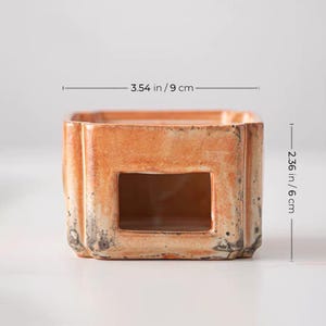 May include: A square, ceramic candle holder with a rectangular opening. The holder is 3.54 inches (9 cm) wide and 2.36 inches (6 cm) tall. It has a distressed, orange-brown finish with dark accents.