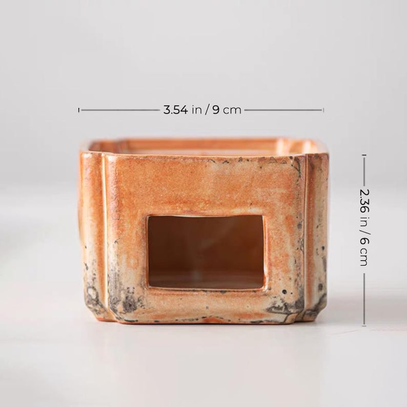 May include: A square, ceramic candle holder with a rectangular opening. The holder is 3.54 inches (9 cm) wide and 2.36 inches (6 cm) tall. It has a distressed, orange-brown finish with dark accents.
