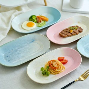 Op de afbeelding: Een assortiment ovale keramische borden in pastelkleuren, waaronder blauw en roze, is gerangschikt op een wit oppervlak. De borden zijn gevuld met verschillende gerechten, zoals eieren, groenten en pasta. Een gouden vork is zichtbaar.