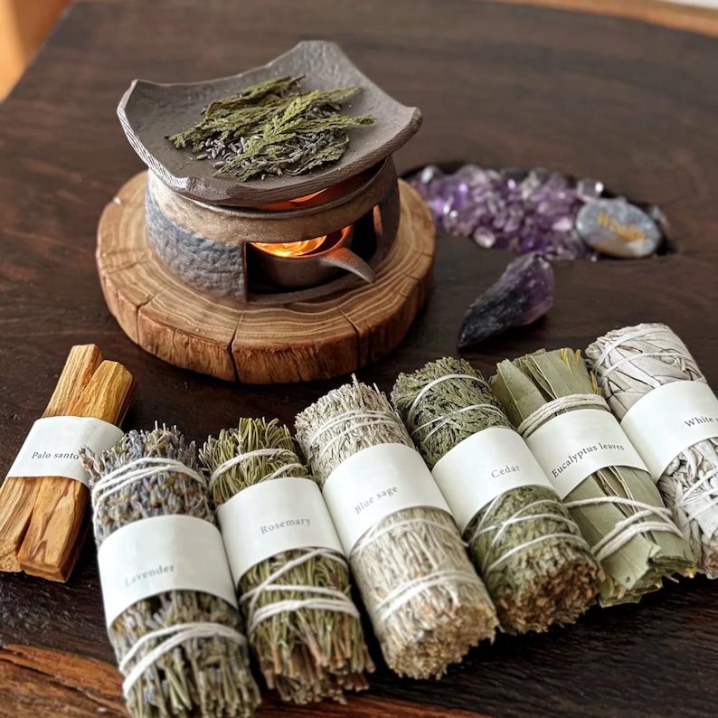 May include: An assortment of aromatherapy items, including bundles of herbs labeled "Lavender", "Rosemary", "Blue sage", "Cedar", "Eucalyptus leaves", and "White sage". Also shown are Palo Santo sticks, a ceramic burner, and crystals.
