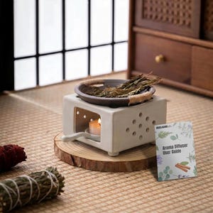 May include: A white ceramic oil burner with a square base and perforated sides, holding a dark brown shallow bowl filled with dried herbs. A lit tea light candle sits inside the base. A small card labeled Aroma Diffuser User Guide is placed nearby.