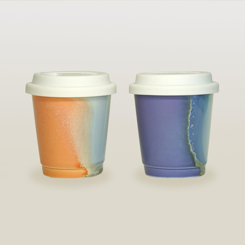 May include: Two ceramic coffee mugs with white lids. The mug on the left is orange and blue, while the mug on the right is purple and blue.