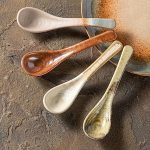 May include: Four ceramic spoons in varying earth tones, including beige, brown, and green, are arranged on a textured surface. The spoons have a long, slender handle and a rounded bowl, with a rustic aesthetic. A ceramic plate is in the background.