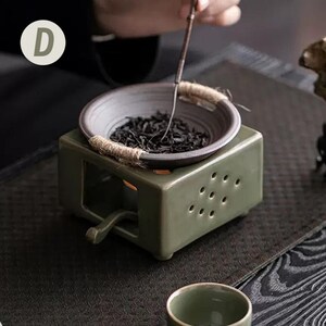 Op de afbeelding: Een groene keramische theeverwarmer met een kleine kom met theebladeren. De kom is omgeven door een touw. De verwarmer heeft een vierkante basis met een handvat en verschillende gaten. Een theegerei is in gebruik.