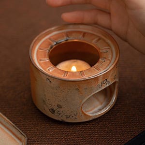May include: A ceramic candle holder with a lit tea light inside. The holder is cylindrical with a circular opening at the top and a side opening. The color is a mix of brown and orange, with a textured surface.