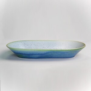 May include: An oval-shaped ceramic serving dish with a gradient of colors. The dish transitions from a light blue interior to a darker blue exterior, with a green rim. The dish has a textured surface.