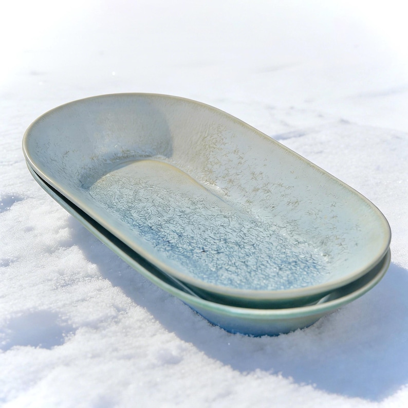 May include: Two oval-shaped ceramic serving dishes. The dishes are stacked, with a light blue and white speckled glaze. The rims have a darker blue hue. The dishes are set against a snowy background.