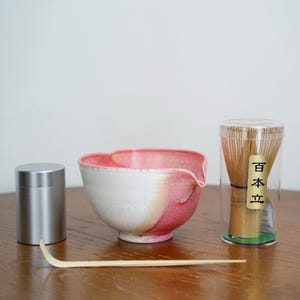 May include: A matcha tea set with a white and pink ceramic bowl, a bamboo whisk in a clear container, a silver container, and a bamboo scoop. The bowl has a unique shape with a spout.