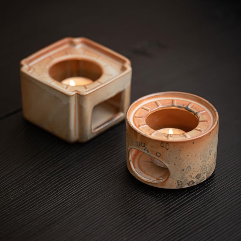 May include: Two ceramic candle holders in a warm, earthy tone. One is hexagonal, the other is round, both with openings for tea light candles. The holders have a rustic, speckled finish and are placed on a dark surface.