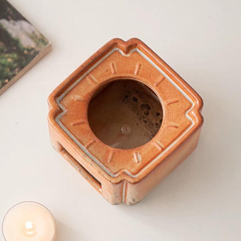 May include: A square, orange ceramic candle holder with a circular opening. The holder has a geometric design with a white trim. The candle holder is sitting on a white surface.