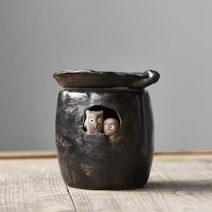 Op de afbeelding: Zwarte keramische plantenbak met een deksel en een kleine opening aan de voorkant. Twee kleine keramische uilen kijken uit de opening.