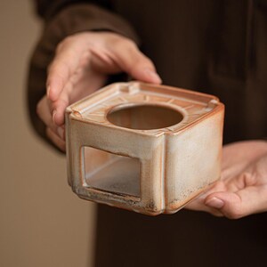May include: A square, ceramic incense burner with a circular opening and a rectangular cutout. The beige and brown tones give it a rustic appearance. The burner is held in a person's hands.