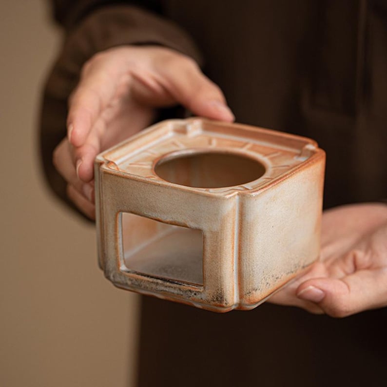 May include: A square, ceramic incense burner with a circular opening and a rectangular cutout. The beige and brown tones give it a rustic appearance. The burner is held in a person's hands.