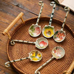 May include: Six ceramic spoons with floral designs. The spoons have a cat-shaped handle and are arranged on a woven tray.