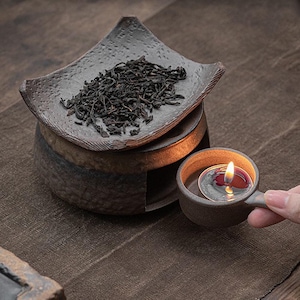 May include: A ceramic tea warmer set with a candle and loose tea. The set includes a small, square dish with tea leaves, a cylindrical base, and a small cup with a lit candle. The items are in shades of brown and gray.