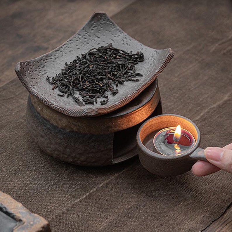 May include: A ceramic tea warmer set with a candle and loose tea. The set includes a small, square dish with tea leaves, a cylindrical base, and a small cup with a lit candle. The items are in shades of brown and gray.