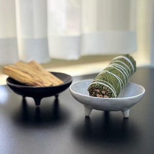 May include: Two small, ceramic bowls with three legs each. One is black and holds several pieces of light brown wood. The other is light gray and contains a bundle of green herbs wrapped with white string. The bowls are on a black surface.