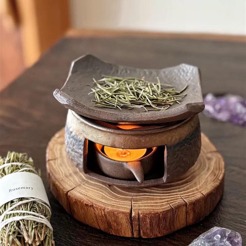 May include: A dark gray ceramic essential oil diffuser with a square top holding dried herbs, a lit candle, and a wooden base. A bundle of rosemary is visible to the left.