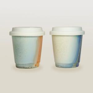 May include: Two ceramic coffee mugs with white lids. The mug on the left has a blue and orange glaze. The mug on the right has a white, blue, and gray glaze.