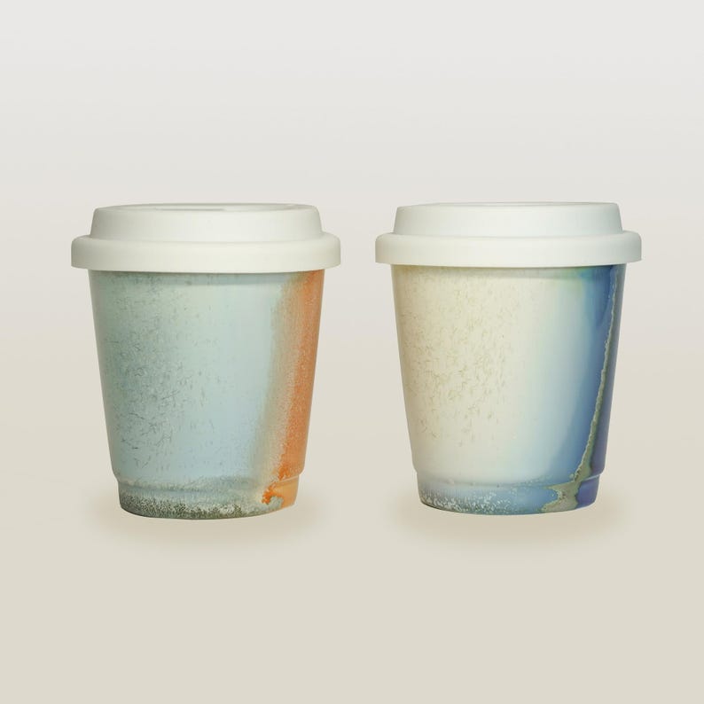 May include: Two ceramic coffee mugs with white lids. The mug on the left has a blue and orange glaze. The mug on the right has a white, blue, and gray glaze.