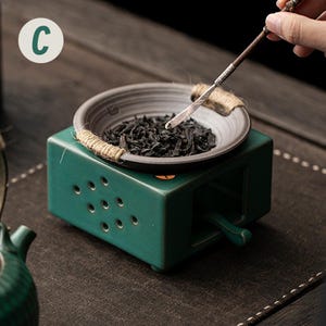 Op de afbeelding: Een teal keramische theeverwarmer met een kleine, ronde, grijze kom gevuld met theebladeren. De kom is omwikkeld met touw en staat bovenop de verwarmer. Een hand houdt een theegerei boven de kom. De letter "C" staat in een witte cirkel linksboven.