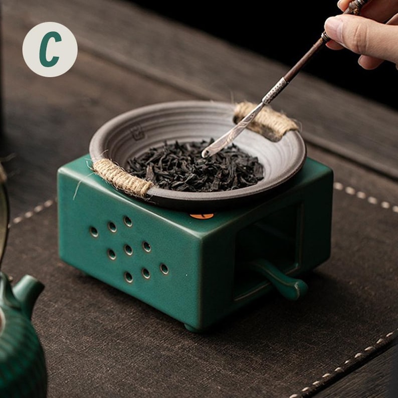 Op de afbeelding: Een teal keramische theeverwarmer met een kleine, ronde, grijze kom gevuld met theebladeren. De kom is omwikkeld met touw en staat bovenop de verwarmer. Een hand houdt een theegerei boven de kom. De letter "C" staat in een witte cirkel linksboven.