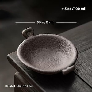 May include: A small, dark brown ceramic bowl with a handle. The bowl is approximately 5.9 inches (15 cm) in diameter and 1.57 inches (4 cm) high, with a capacity of about 3 oz (100 ml). The bowl has a textured surface and is designed for serving.